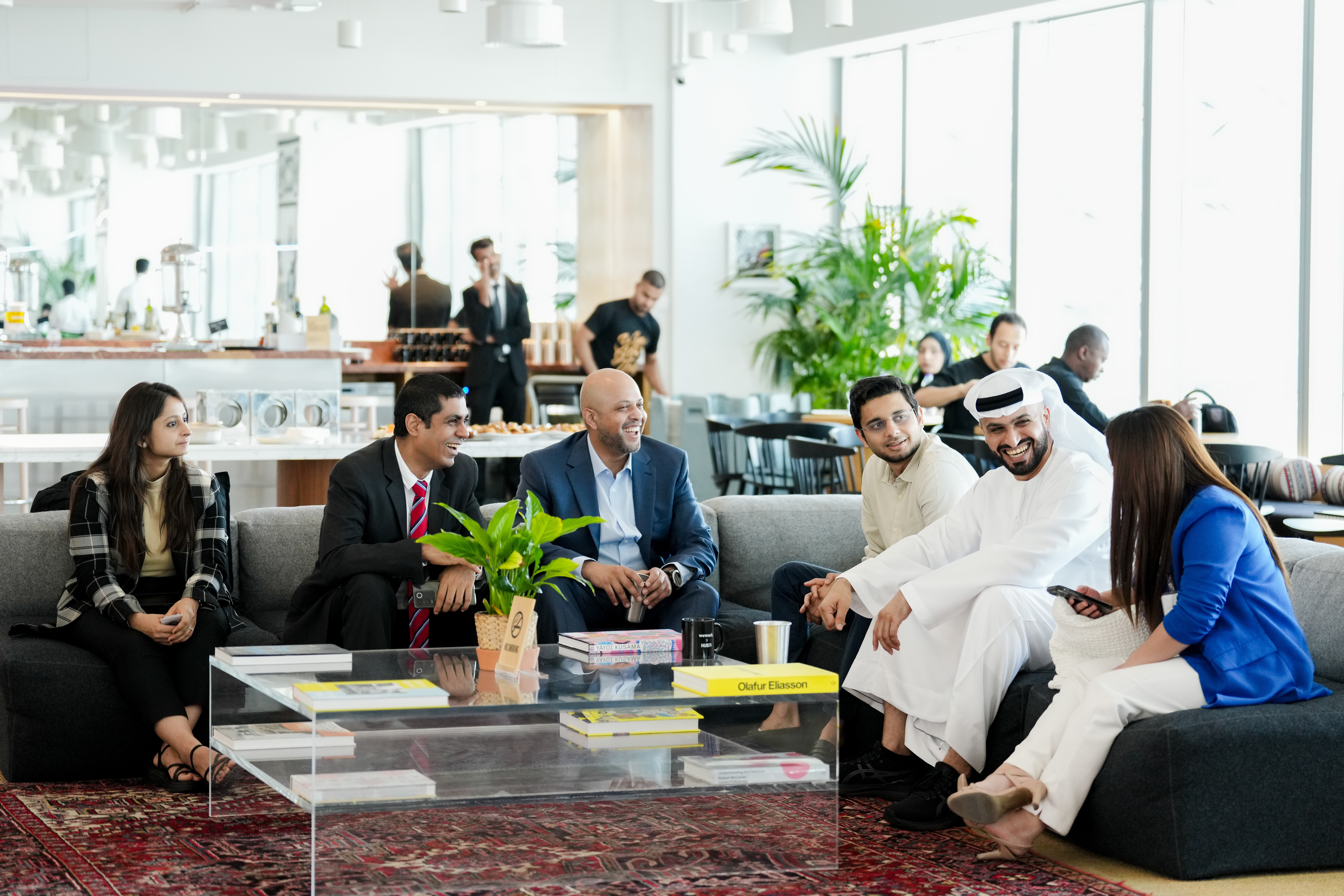 An image of business men and women dressed smartly sat on sofas in a restaurant with people behind them and a coffee table in front.