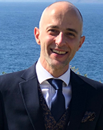 Ioannis Kokkoris wearing a suit and blue tie, standing in front of the ocean