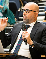 Noam wears dark glasses dressed in a dark suit holding a microphone