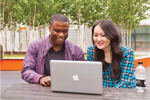 two students sitting looking at a laptop, outside.