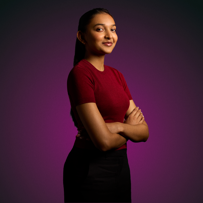 A female student wearing a red top and black trousers with arms crossed smiling looking forward and smiling, with a purple background.